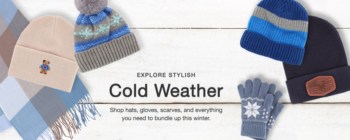 A flat-lay winter apparel collection on a white wood background featuring a beige knit beanie with a small embroidered character, a gray and blue striped beanie with a pom-pom, a blue and gray striped beanie, a navy beanie with a brown patch, a pair of blue and gray gloves with a snowflake, and a plaid scarf.